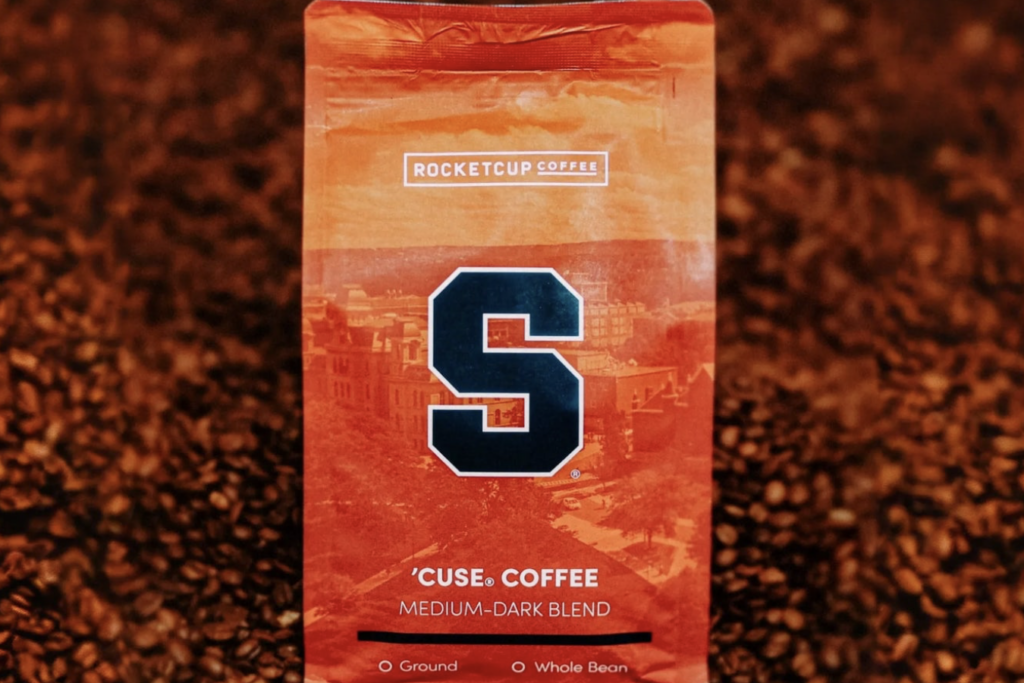 A orange RocketCup Coffee bag with the Syracuse University logo sits in a display of thousands of coffee beans