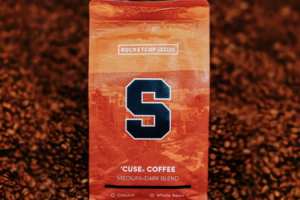 A orange RocketCup Coffee bag with the Syracuse University logo sits in a display of thousands of coffee beans