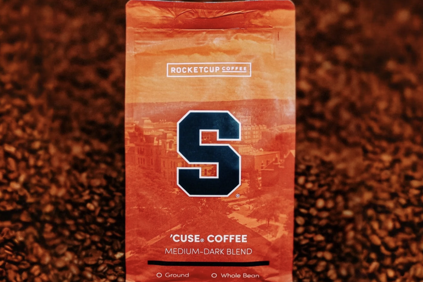 A orange RocketCup Coffee bag with the Syracuse University logo sits in a display of thousands of coffee beans