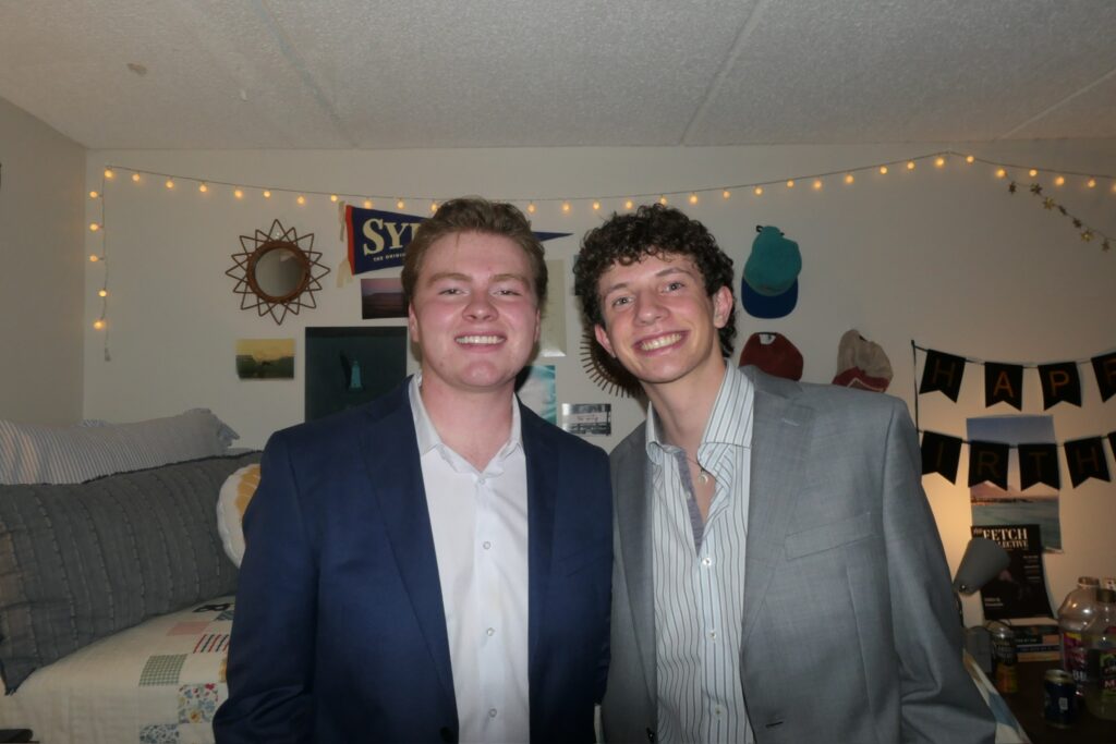 Syracuse University students and influencers Spencer Howard and Josh Spina stand in a dorm room together dressed in suits.