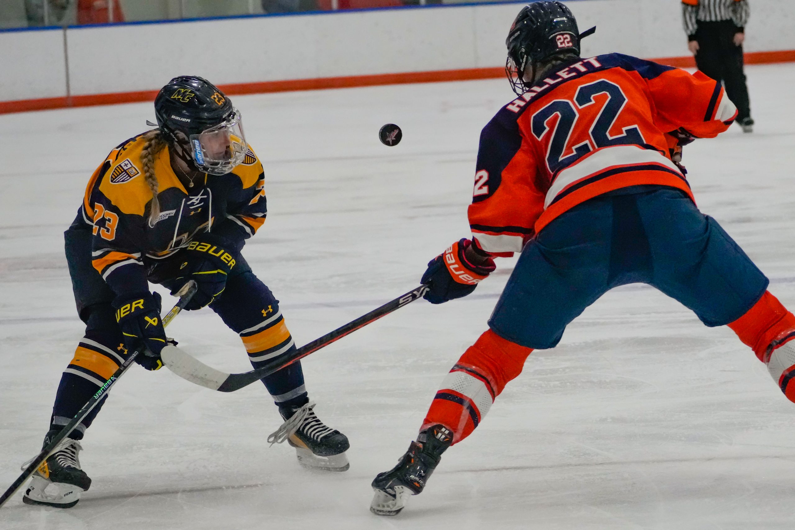 The puck gets knocked up as Merrimack's Hannah Corneliusen faces defender Charlotte Hallett (#22).