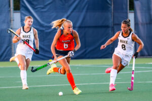 Field Hockey player in orange jersey runs between two other players in white jerseys