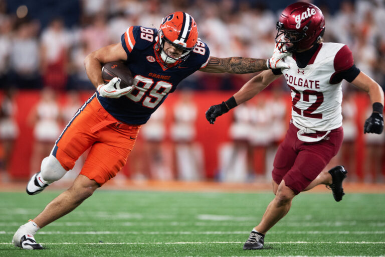 Football player in blue and orange stiff arms opposing football player in white and maroon
