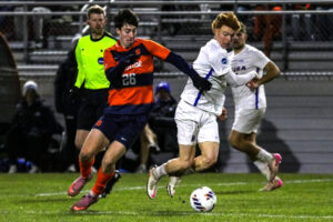 Male soccer player in orange fights for the ball against a male soccer player in white on a grass field while a ref in neon green watches.