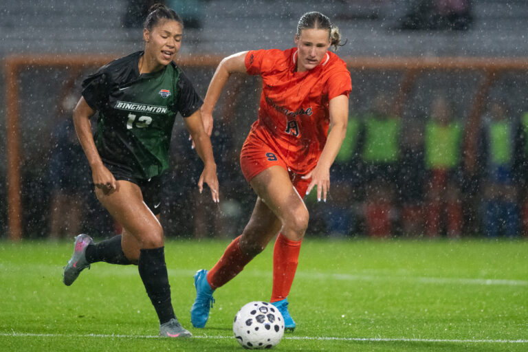 Woman in orange jersey fights for the soccer ball against woman in green jersey in the rain.