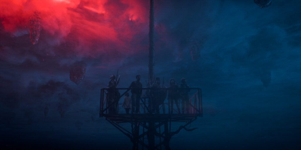 In Stranger Things Season 5, the main characters stand on top of a tall radio tower with the blue and red background of the Upside Down behind them.