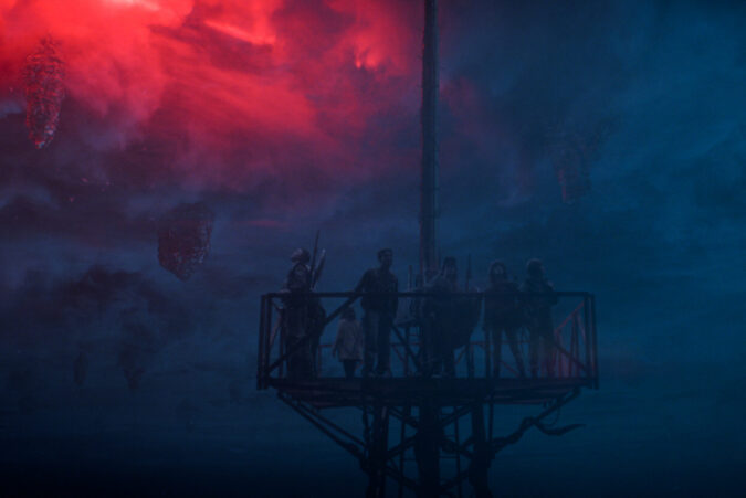 In Stranger Things Season 5, the main characters stand on top of a tall radio tower with the blue and red background of the Upside Down behind them.