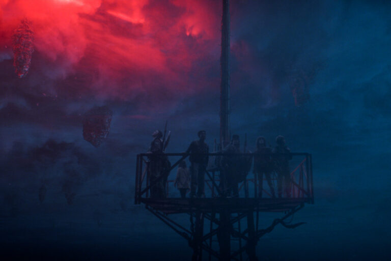 In Stranger Things Season 5, the main characters stand on top of a tall radio tower with the blue and red background of the Upside Down behind them.