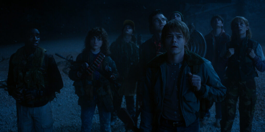 In Stranger Things Season 5 a group of characters stand dressed in tactical gear and with weapons in the dark background of the Upside Down.