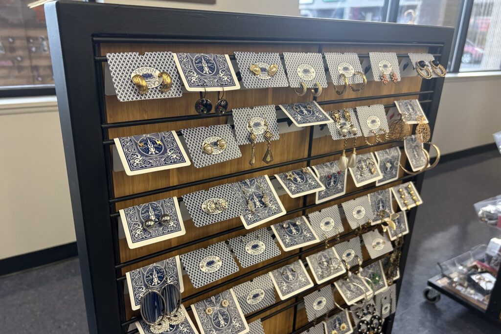 On a wooden rack, a series of blue bicycle playing cards sit horizontally; hanging from them each are pairs of colorful earrings.
