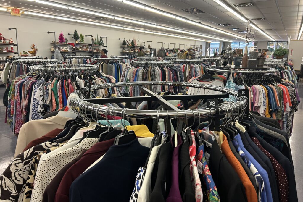 A series of clothing racks in a thrift store, from which range multiple pieces of clothing in all colors.