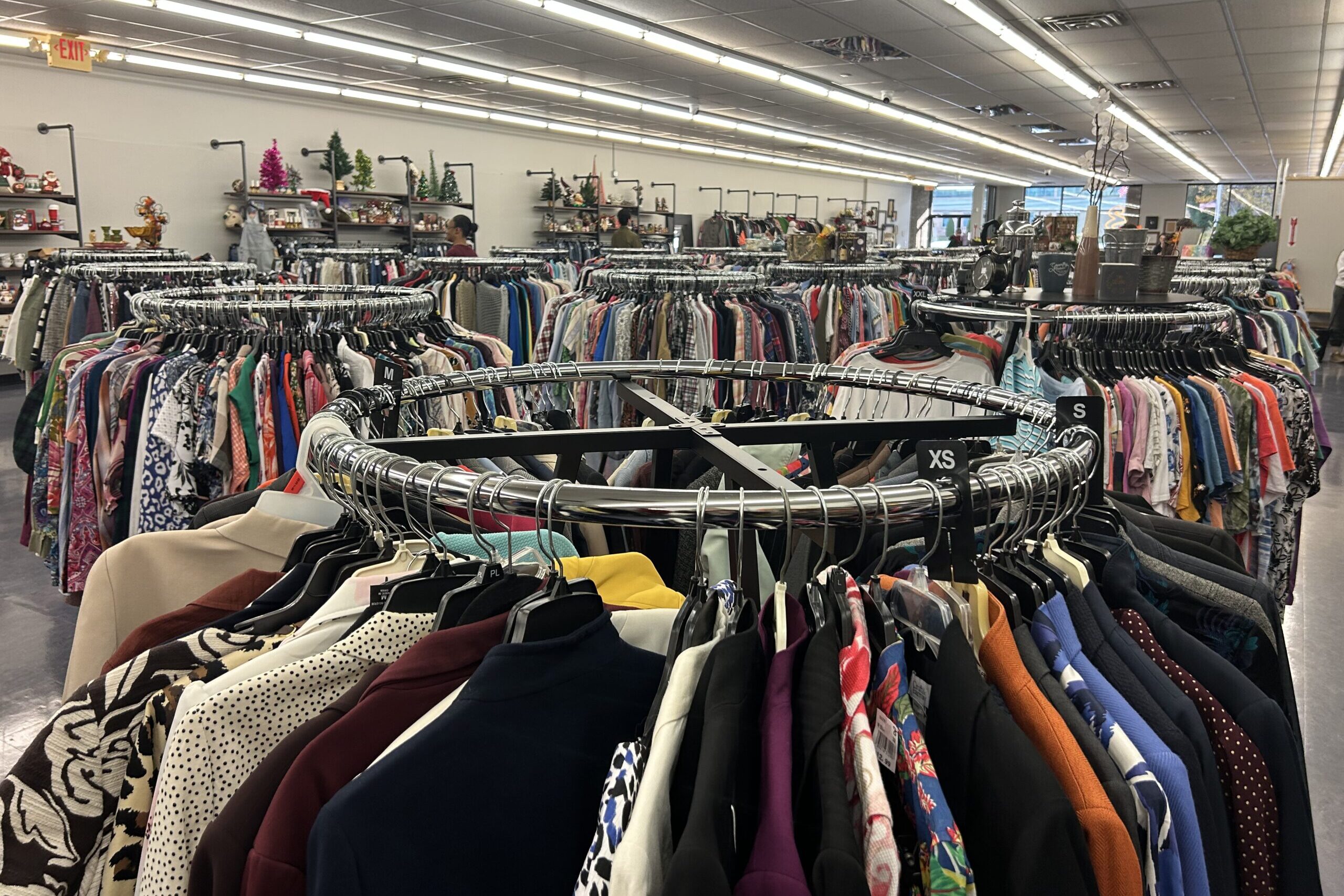 A series of clothing racks in a thrift store, from which range multiple pieces of clothing in all colors.