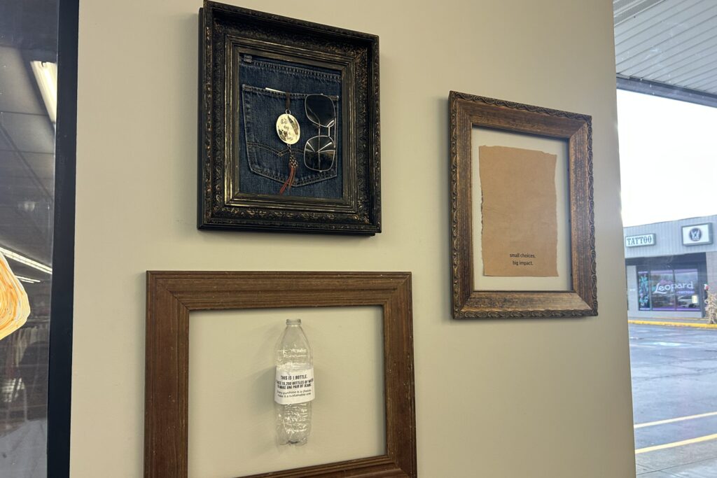 Three photo frames hang on a beige wall. In the top frame is a square of denim jeans, in the middle frame is a paper bag and the bottom frame holds a small plastic water bottle.