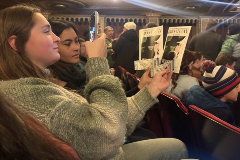 In the Landmark Theater, two young women hold up their "Wicked" programs and tickets in front of the stage to take a picture.