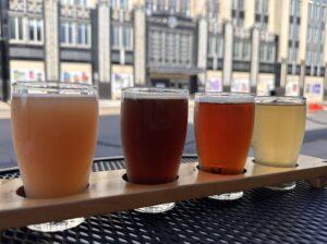 Four drinks from Talking Cursive Brewery (left to right: orange colored, brown, red and yellow) are arranged in a drink carrier on a metal table.