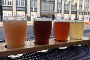 Four drinks from Talking Cursive Brewery (left to right: orange colored, brown, red and yellow) are arranged in a drink carrier on a metal table.