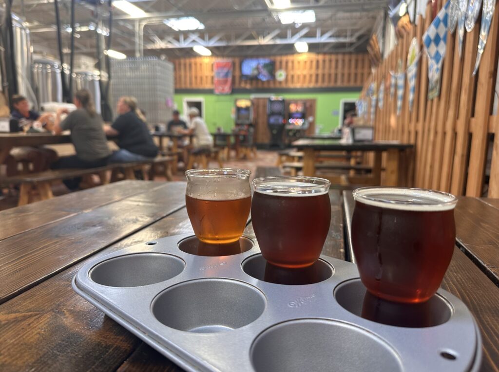 Three drinks from Willow Rock Brewery are served in a metal cupcake pan. In the background patrons sit at long wooden tables.