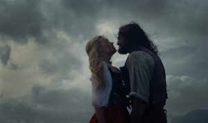 In "Wuthering Heights" against a grey sky, a woman with blonde hair and a shaggy man stand close to each other.