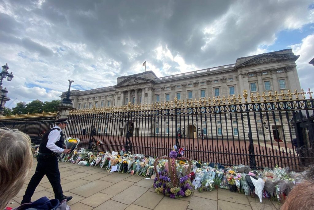 Buckingham Palace with flowers lined up against the fence, and a guard shown carrying another bouquet over towards the fence.