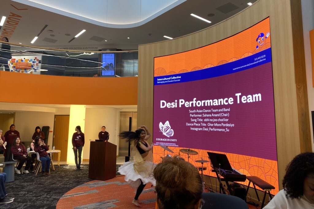 A dancer performs a Desi dance in front of a screen for students and faculty in an auditorium.
