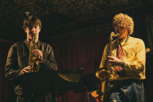 two saxophone players performing in dim lighting at jazz club