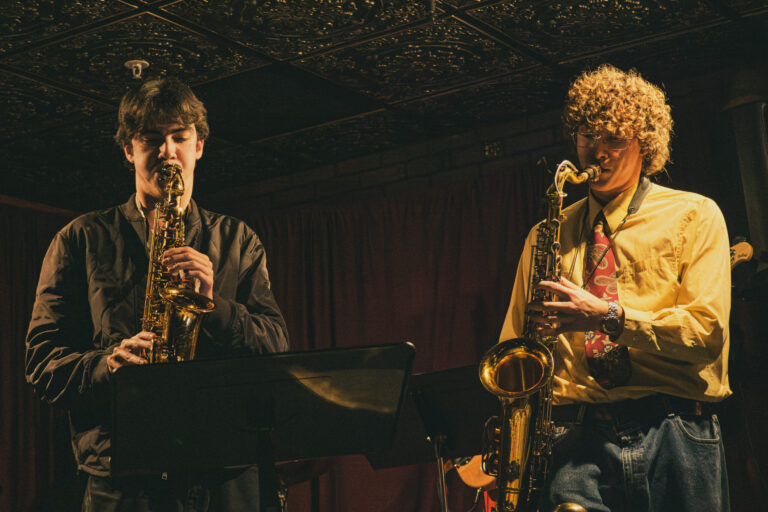 two saxophone players performing in dim lighting at jazz club