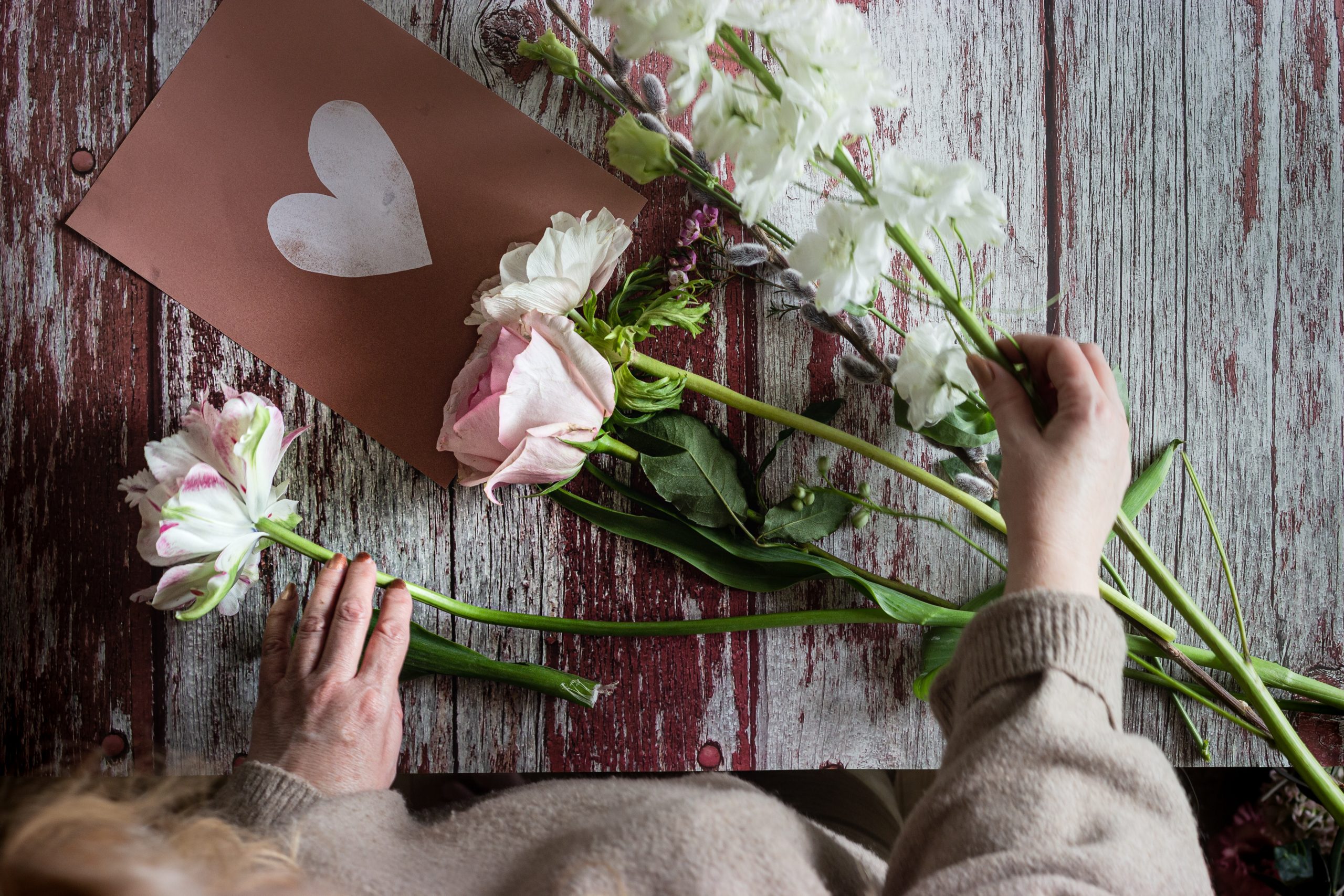 An individual places flowers around a handmade card with a heart drawn on it.