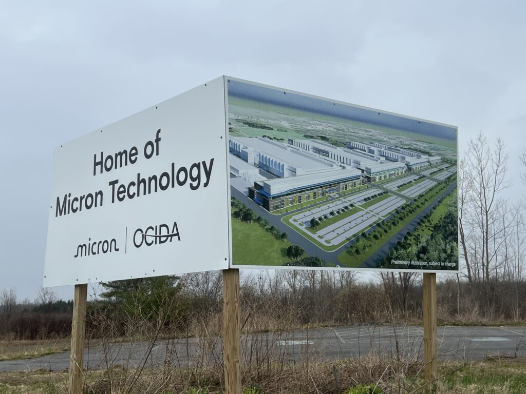 A sign of a microchip complex that stands on the side of a road in rainy weather next to trees.