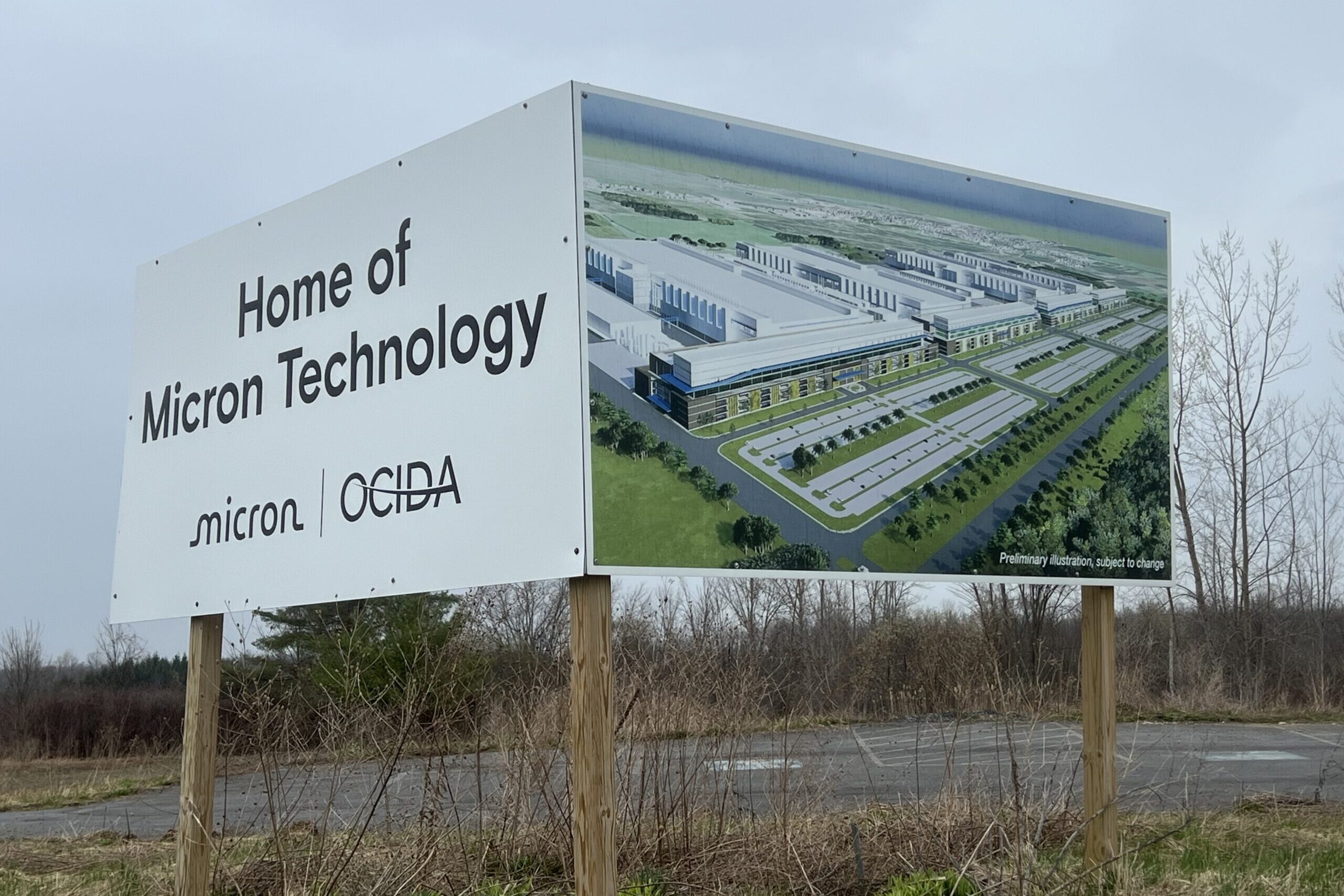 A sign of a microchip complex that stands on the side of a road in rainy weather next to trees.